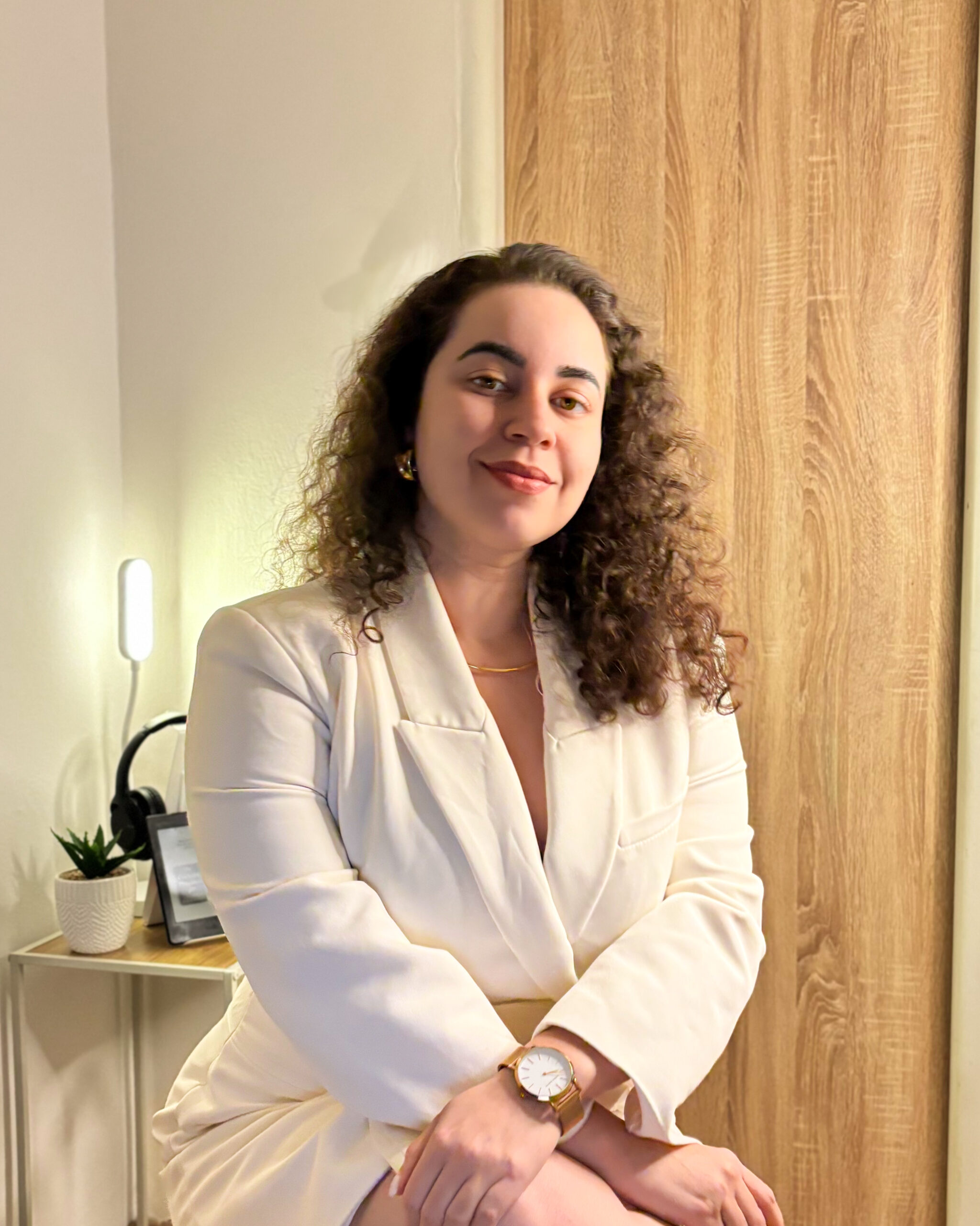 A woman with curly hair wearing a white blazer and skirt sits in a well-lit room. They are accessorized with a gold watch and necklace. The background includes a wooden door, a small table with a potted plant, headphones, and a lit lamp.